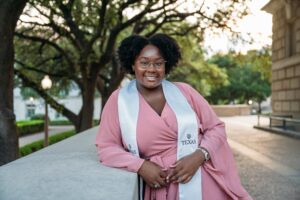 Photo of a black woman with a pink dress wearing a white UT graduation stole. Jordan is leaving against a stone wall with trees in the background