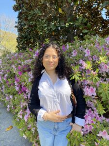Photo of Latina woman with black curly hair and classes wearing a white button up shirt, black sweater and jeans in front of a hedge of purple flowers