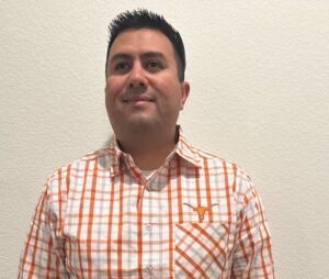A person smiling with straight black hair, wearing a burnt orange and white checkered dress shirt. Above the pocket of the shirt appears to be the University of Texas Longhorn logo. He is also posing in front of a white wall.